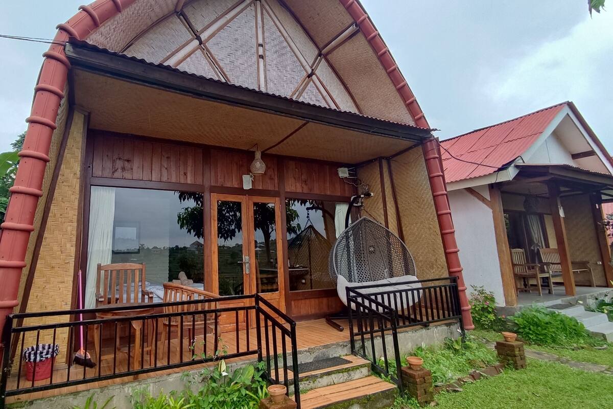Una casa de madera con techo inclinado, puerta de vidrio, porche con barandilla negra y una silla colgante.