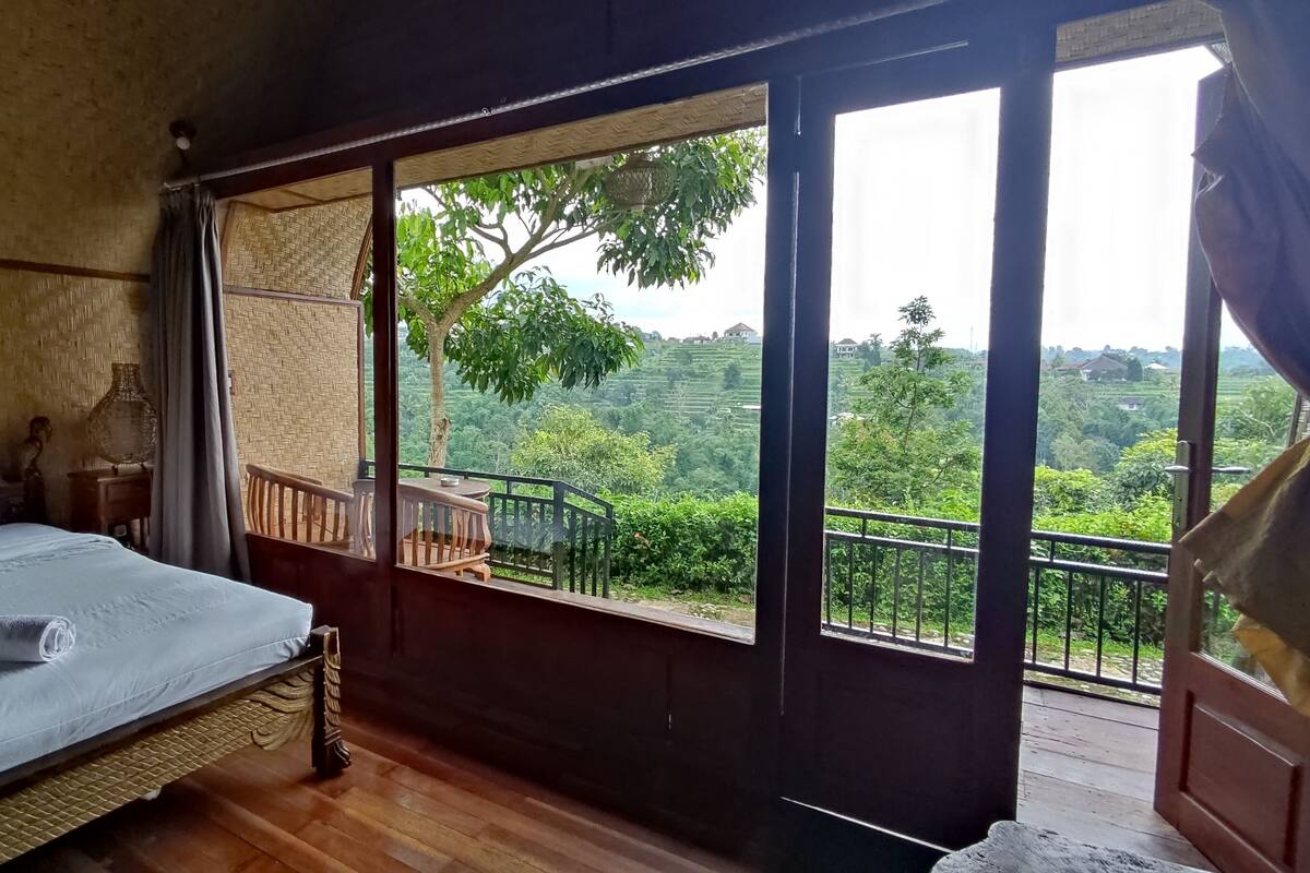 Un dormitorio con una cama grande, piso de madera, balcón con barandilla y vista a un paisaje exuberante.