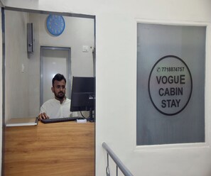 A man sitting at a desk with a computer, a clock on the wall, and a sign for 'Vogue Cabin Stay' with a phone number. - Vogue Cabin Stay Near NESCO (Mumbai)