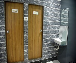 Two wooden doors with "TOILET" signs, a marble-patterned wall, and a small sink with a mirror. - Vogue Cabin Stay Near NESCO (Mumbai)