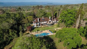 Una casa grande de dos pisos con piscina, rodeada de un bosque denso y montañas al fondo.