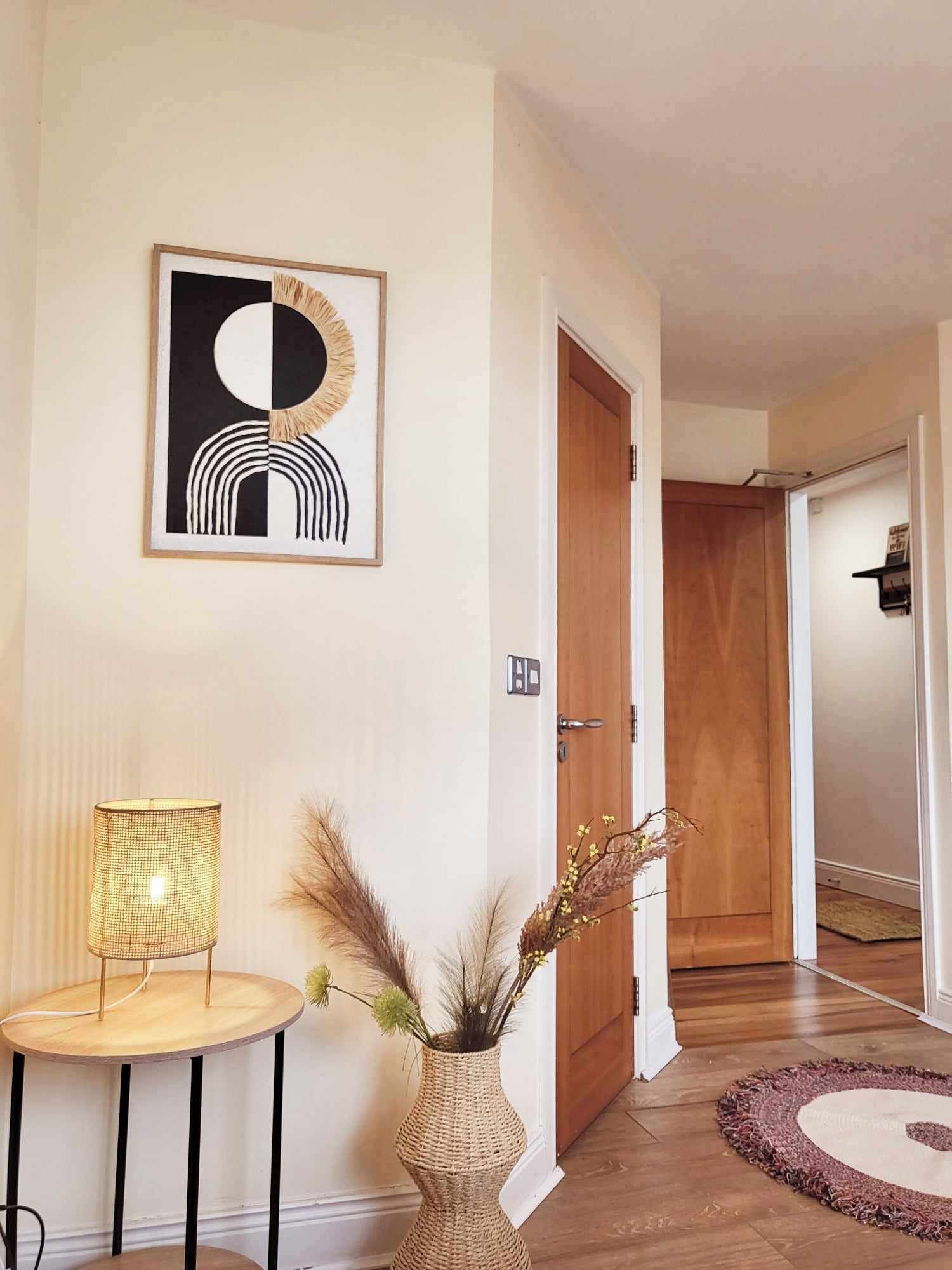 A hallway with a wooden door, a framed abstract artwork, a small round table with a lamp, and a vase with dried plants.
