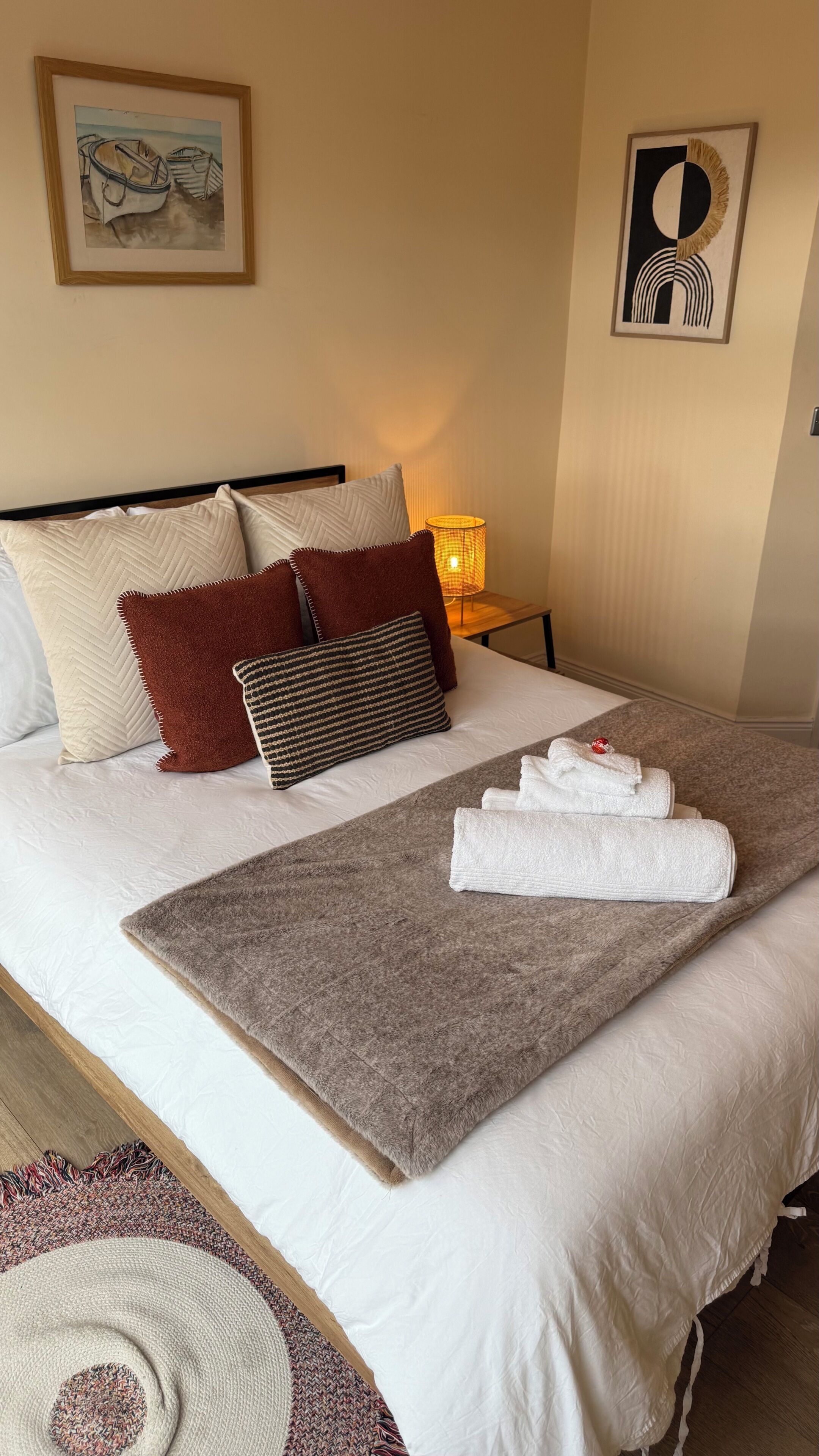 A neatly made bed with pillows, a blanket, and towels. There are two wall-mounted artworks, a lamp, and a wooden headboard.