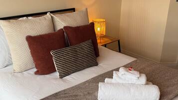 A neatly made bed with pillows, a blanket, and towels. There are two wall-mounted artworks, a lamp, and a wooden headboard.