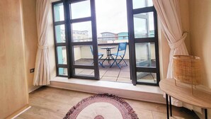 A room with a large window, a small balcony with a table and chairs, and a wicker basket on a side table. - Panoramic Two-bedroom Penthouse (Cork)