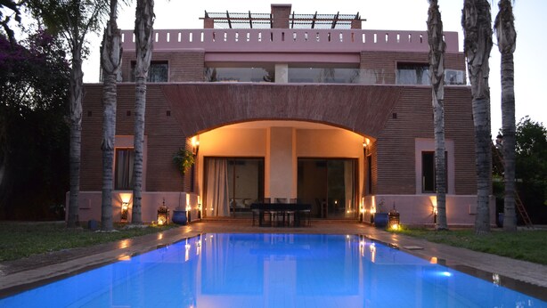 A swimming pool with a covered area, palm trees, and a building with a flat roof and large windows. - Villa in the Heart of Marrakech (Marrakech)