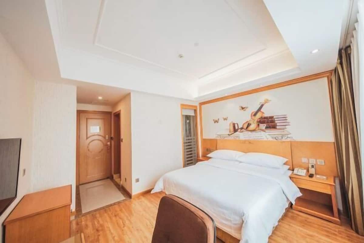 A hotel room with a bed, a desk, a chair, a TV, and a wall with a guitar and books.