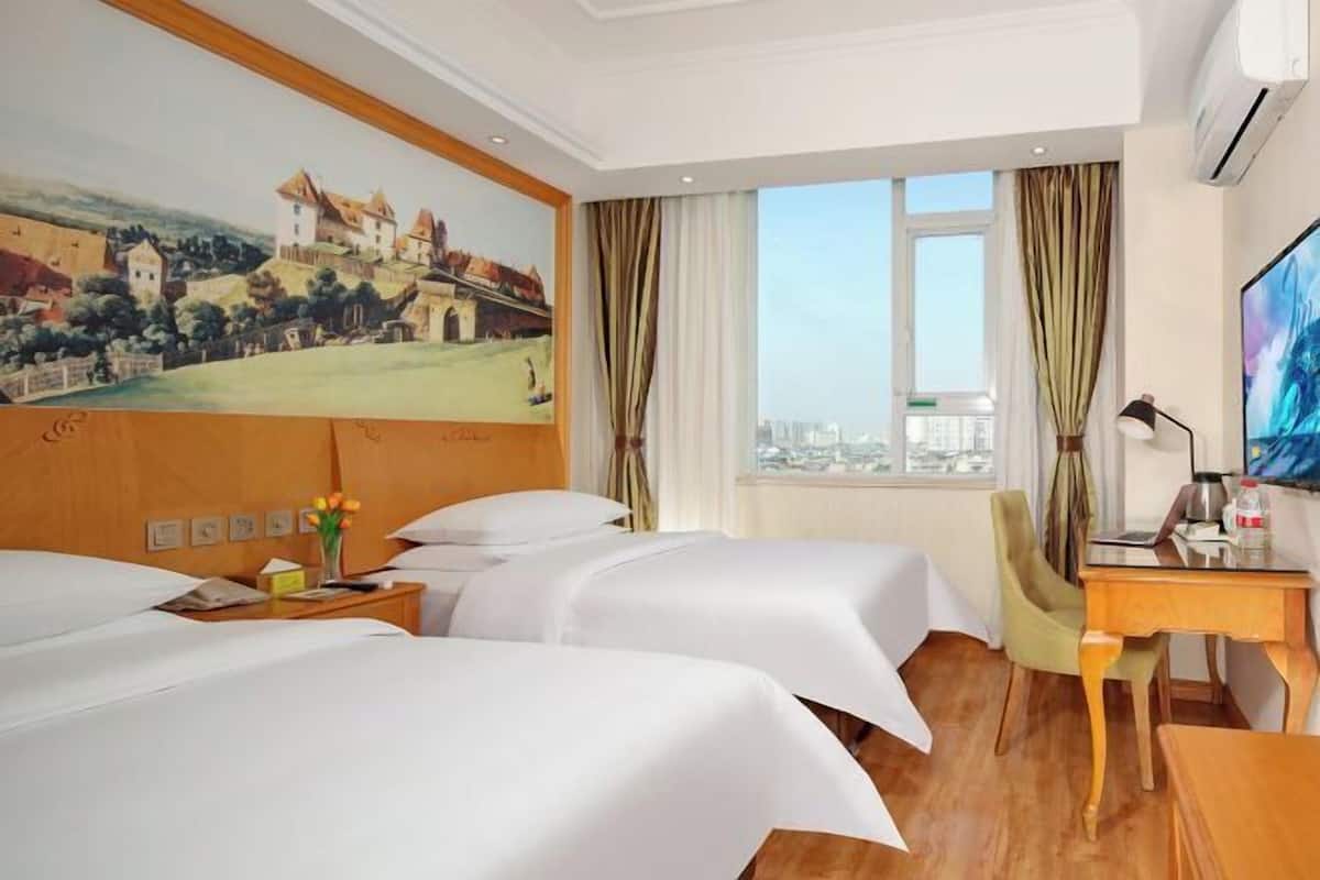 A hotel room with two beds, a desk, a chair, a painting on the wall, and a window with curtains.