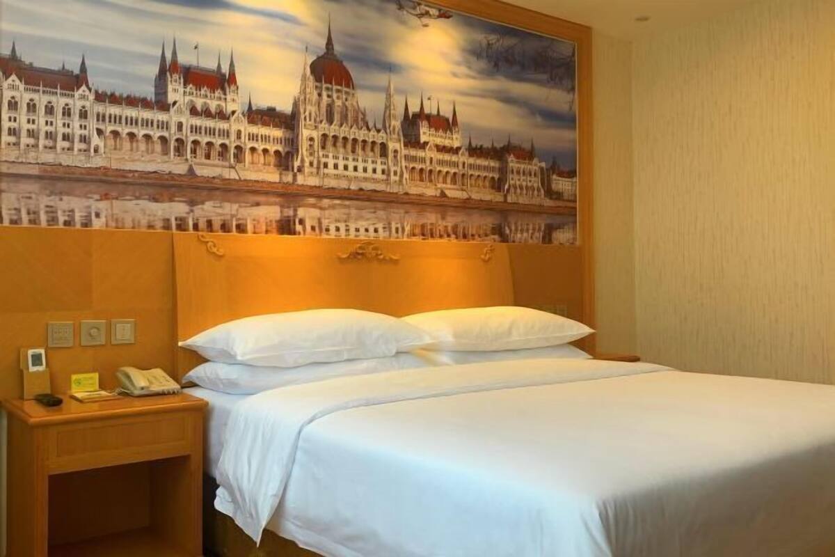A hotel room with a large bed, a wooden headboard, a nightstand with a phone, and a painting of a building on the wall.