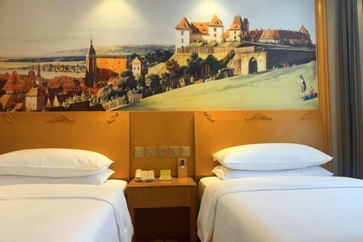 A hotel room with two beds, a wooden headboard, a nightstand with a phone and a small table, and a painting of a castle on the wall.