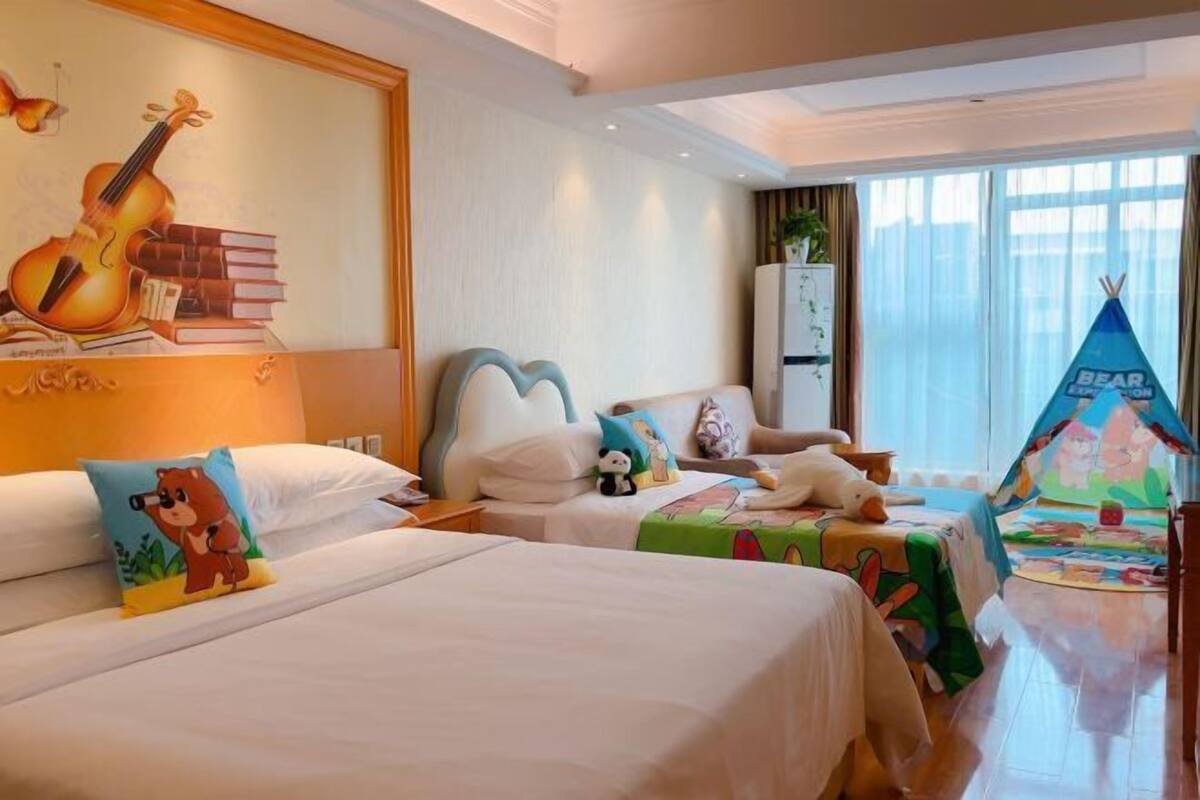 A hotel room with two beds, a large wall mural, a bear-themed pillow, a tent-like decoration, and a window with curtains.