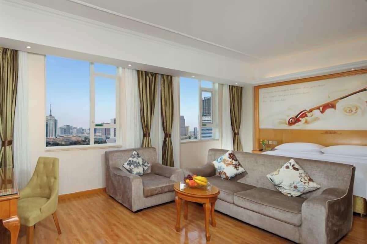 A hotel room with two sofas, a small table with fruit, a bed, and a large window with curtains.