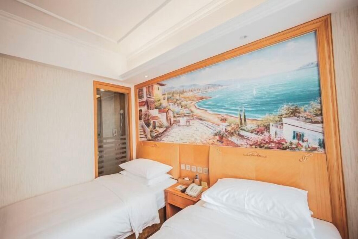 A hotel room with two beds, a painting of a coastal scene, and a wooden headboard.