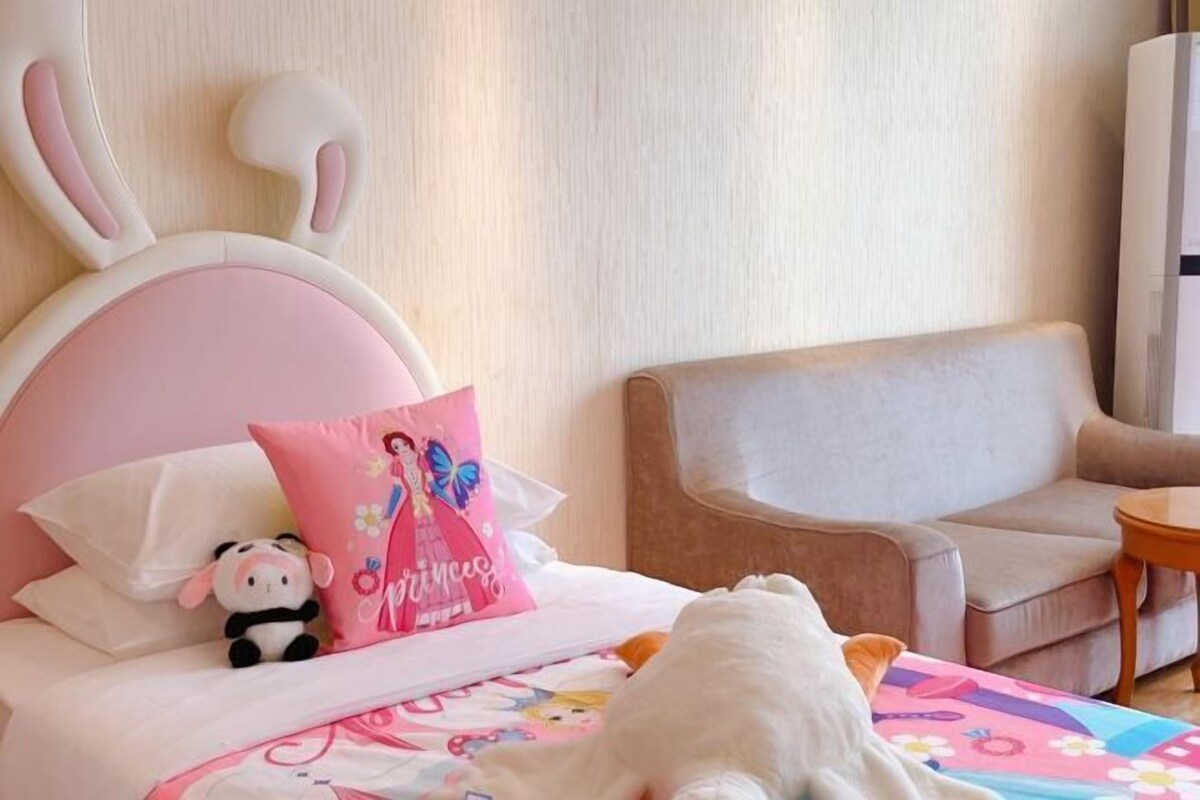 A hotel room with a bed, a plush toy, a pink pillow, and a chair.