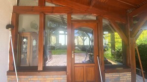 A wooden porch with a glass door and windows, brick base, and a view of a garden and outdoor seating area. - Villa L'estancade Domaine du Douc Tourisme Dans une Ferme de la Foret des Landes (liposthey)