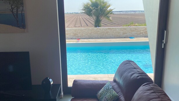 A living room with a brown leather sofa, a television, and a view of a swimming pool through an open door. - Villa L'estancade Domaine du Douc Tourisme Dans une Ferme de la Foret des Landes (liposthey)
