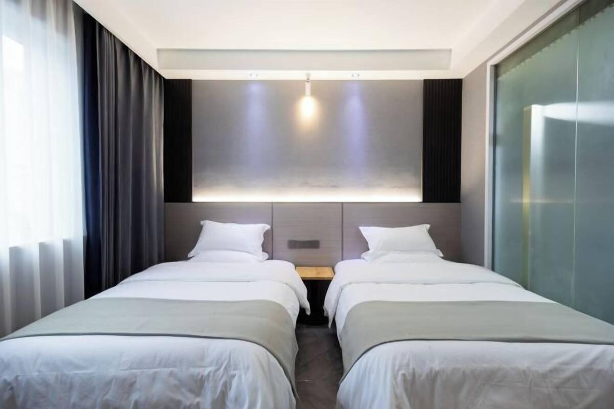 Habitación de hotel con dos camas, un ventanal grande con cortinas, una moderna cabecera y una lámpara de pared.