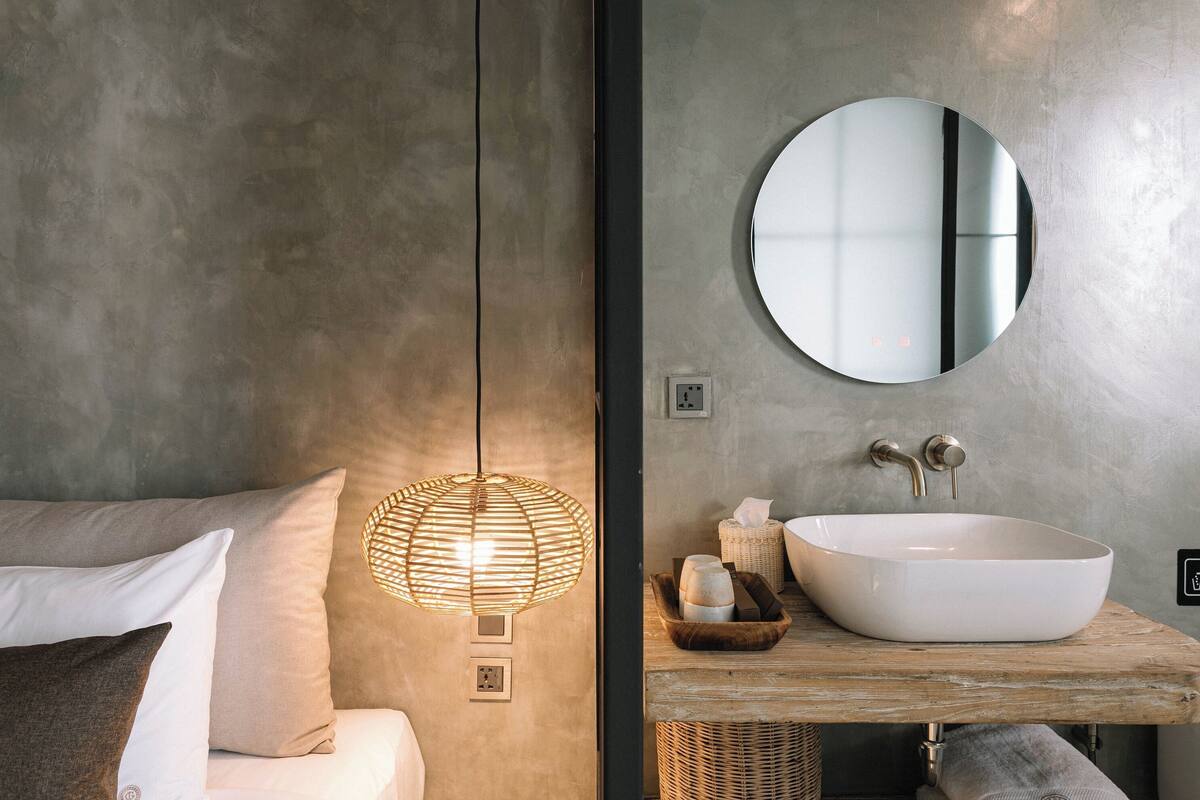 Une salle de bain moderne avec un grand miroir rond, un lavabo sur colonne blanc, un meuble-lavabo en bois et un luminaire suspendu.