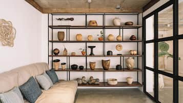 A room with a beige sofa, patterned pillows, and a wall-mounted shelf with various pottery and decorative items.