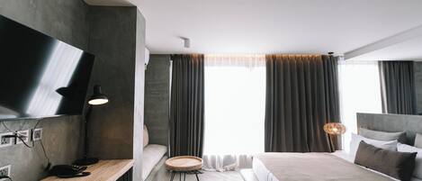 Quarto de hotel moderno com uma cama grande, uma escrivaninha com televisão de tela plana, uma pequena mesa redonda e uma grande janela com cortinas.