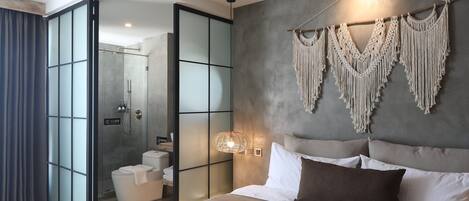 A modern bedroom with a large bed, a macrame wall hanging, and a sliding glass door leading to a bathroom.