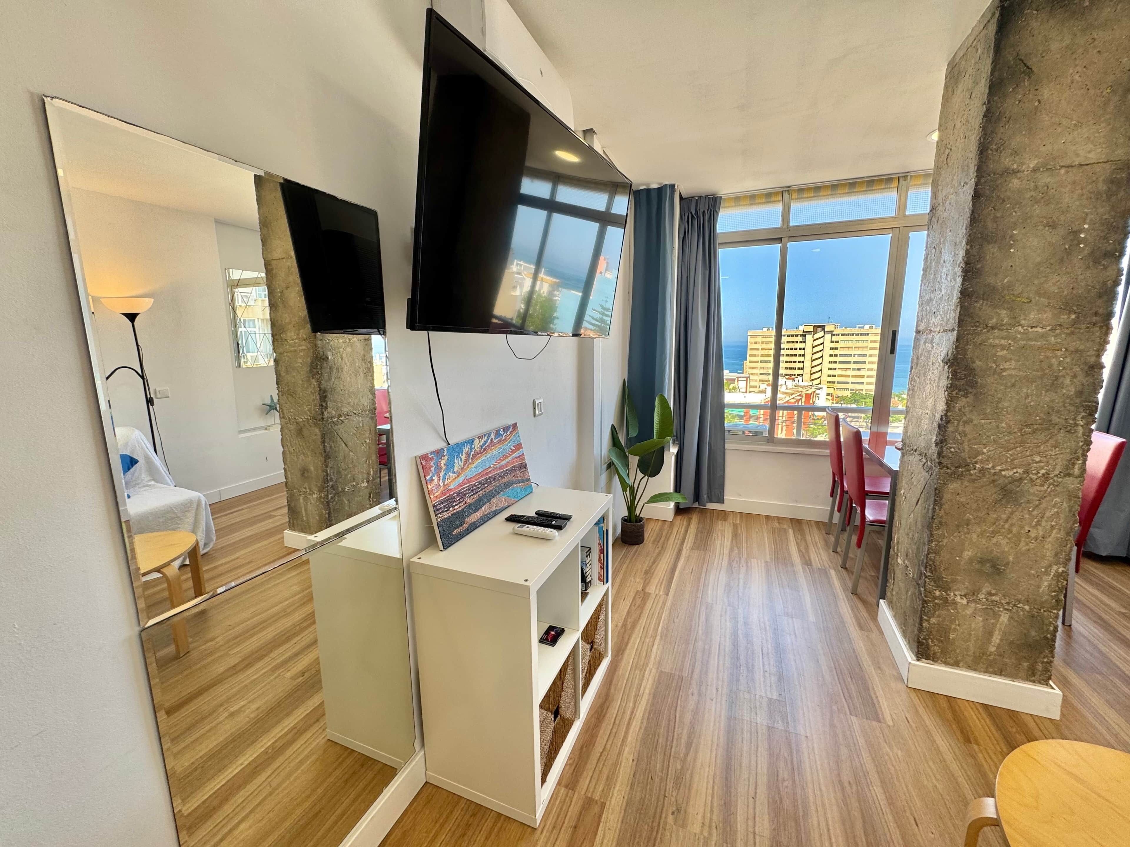 Una habitación de hotel moderna con un espejo grande, un televisor de pantalla plana, una pequeña mesa con una planta y vistas a la ciudad a través de amplias ventanas.