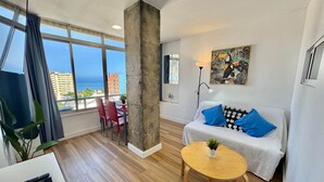 A modern living room with a large window, a sofa with patterned cushions, a coffee table, and a dining area with chairs. - Paraty de SteraM Flats Torremolinos (Torremolinos)