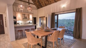 Een eetgedeelte met een houten tafel en rieten stoelen, een keuken met moderne apparatuur, en een groot raam met uitzicht op de omgeving.