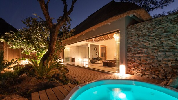 A hot tub in a wooden deck area with a stone wall and a glass-enclosed room with a thatched roof.