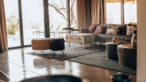 A spacious living room with a high ceiling, large windows, and a view of trees. There is a sofa, a coffee table, and a rug. - Villa Scarabee: Wonderful luxury villa in the middle of nature (Hoedspruit)