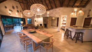 Amplia sala de estar con una larga mesa de madera, múltiples sillas y una chimenea de piedra. Hay luces colgantes y una zona de cocina con taburetes.