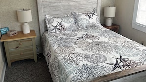A bedroom with a wooden bed frame, a patterned bedspread, two bedside lamps, a nightstand with a lamp, and three wall art pieces depicting ocean scenes. - Historic Harkers Island Bungalow (Harkers Island)