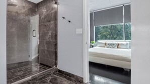 Un bagno moderno con cabina doccia in vetro, pavimento in marmo e vista su una camera da letto con un letto e una finestra.