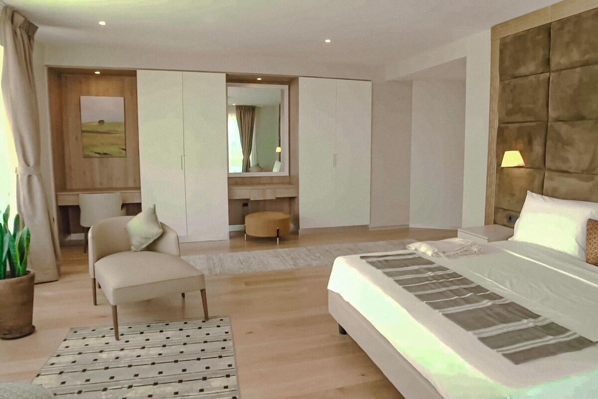 A modern hotel room with a large bed, a sitting area with a chair, a desk, and a mirror.