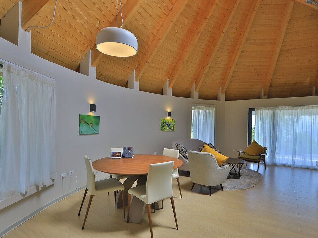 A spacious room with a high wooden ceiling, a round wooden dining table with chairs, a sofa with cushions, and a small side table.