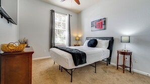 Ein Schlafzimmer mit Bett, Deckenventilator, Fenster mit Vorhängen, Nachttisch mit Lampe und einem Wandbild.