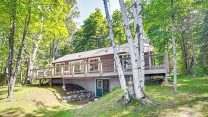 A two-story wooden cabin with a deck, surrounded by trees and a grassy area. - Quiet Presque Isle Home w/ Grill: Families Welcome (Presque Isle)