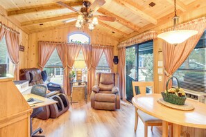L’intérieur chaleureux d’un chalet avec un plafond en bois, un ventilateur de plafond, un fauteuil massant, un bureau avec une chaise, une table à manger avec un panier, et de grandes fenêtres avec des rideaux.