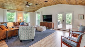 A spacious living room with a wooden ceiling, a television, a sofa, a coffee table, and a large window providing a view of the outdoors. - Views on 12 Acres: Ozark Mountain Cabin! (Parthenon)
