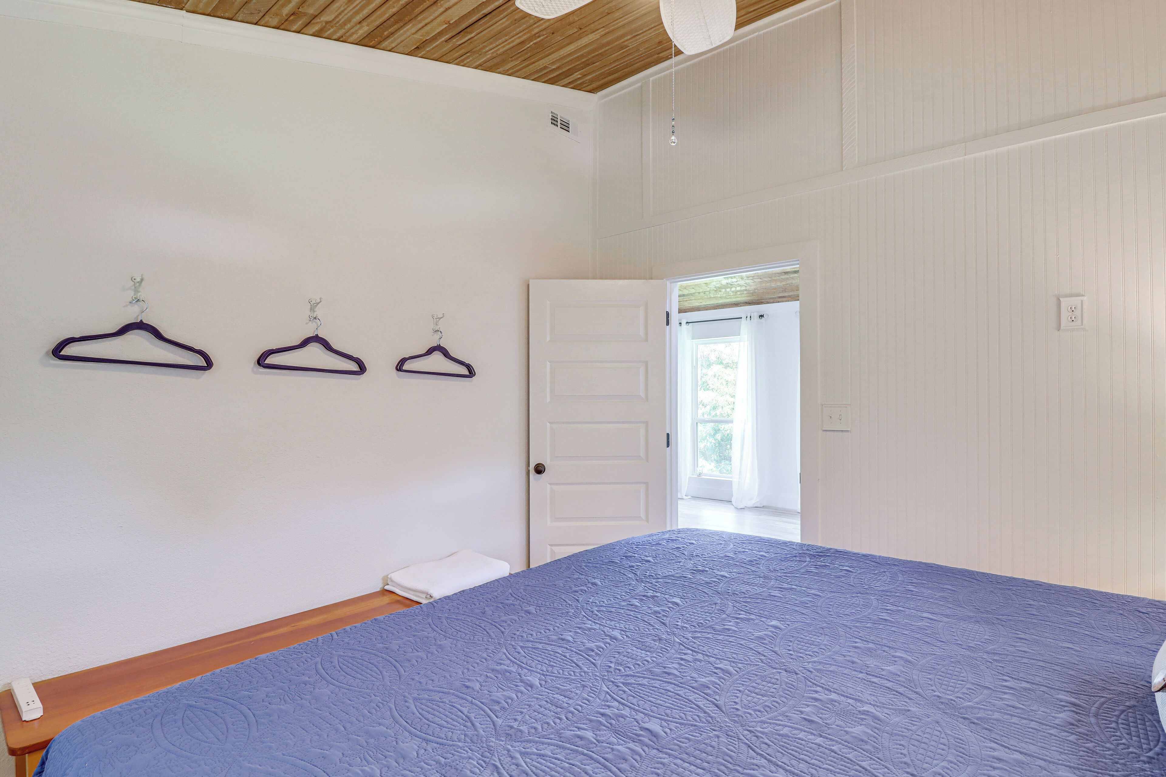 A bedroom with a large bed, wooden ceiling, and white walls. There are three purple hangers on the wall and a white door leading to another room.
