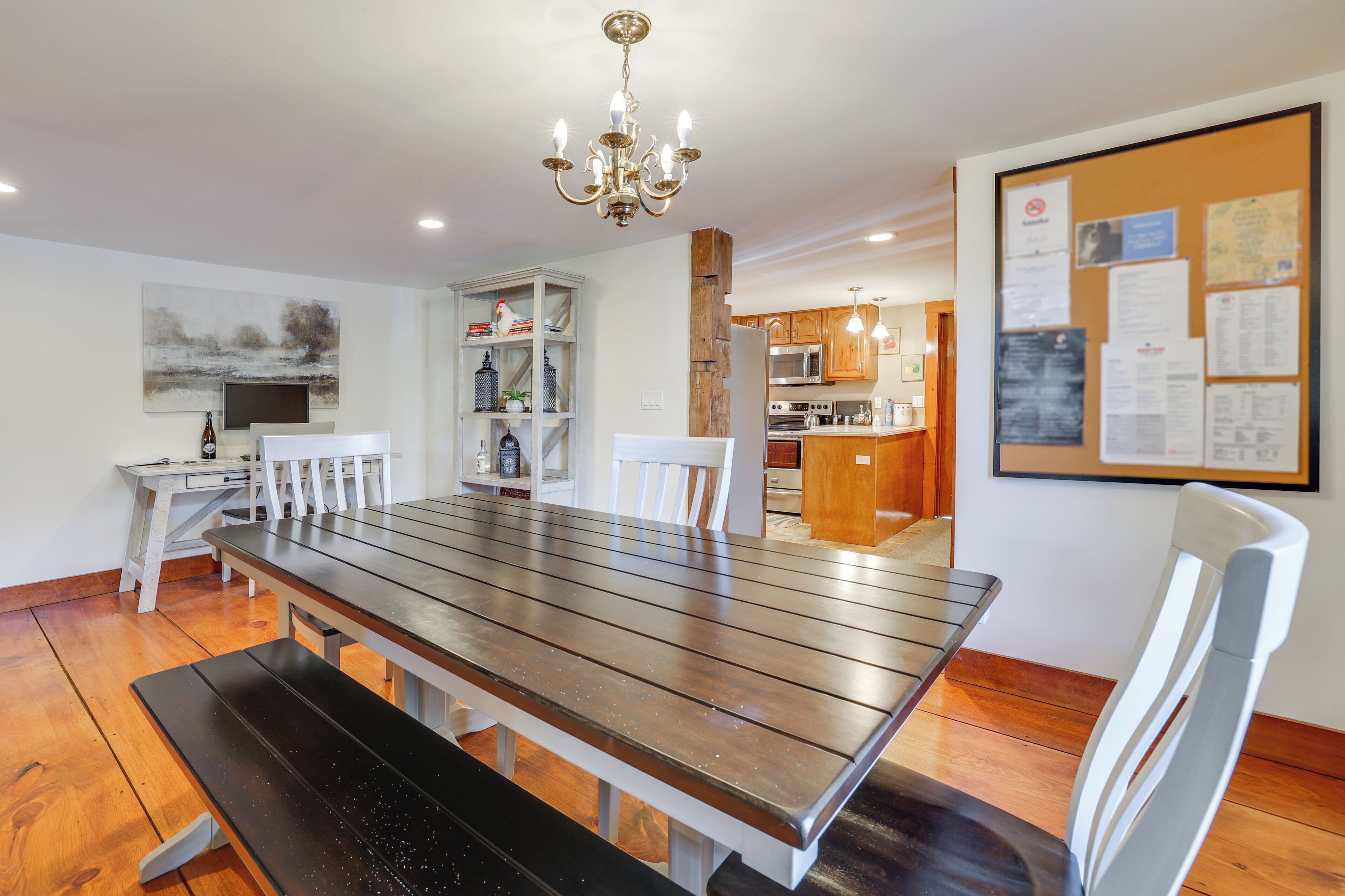 A dining area with a wooden table, white chairs, a bench, a chandelier, a whiteboard with papers, and a kitchen in the background.