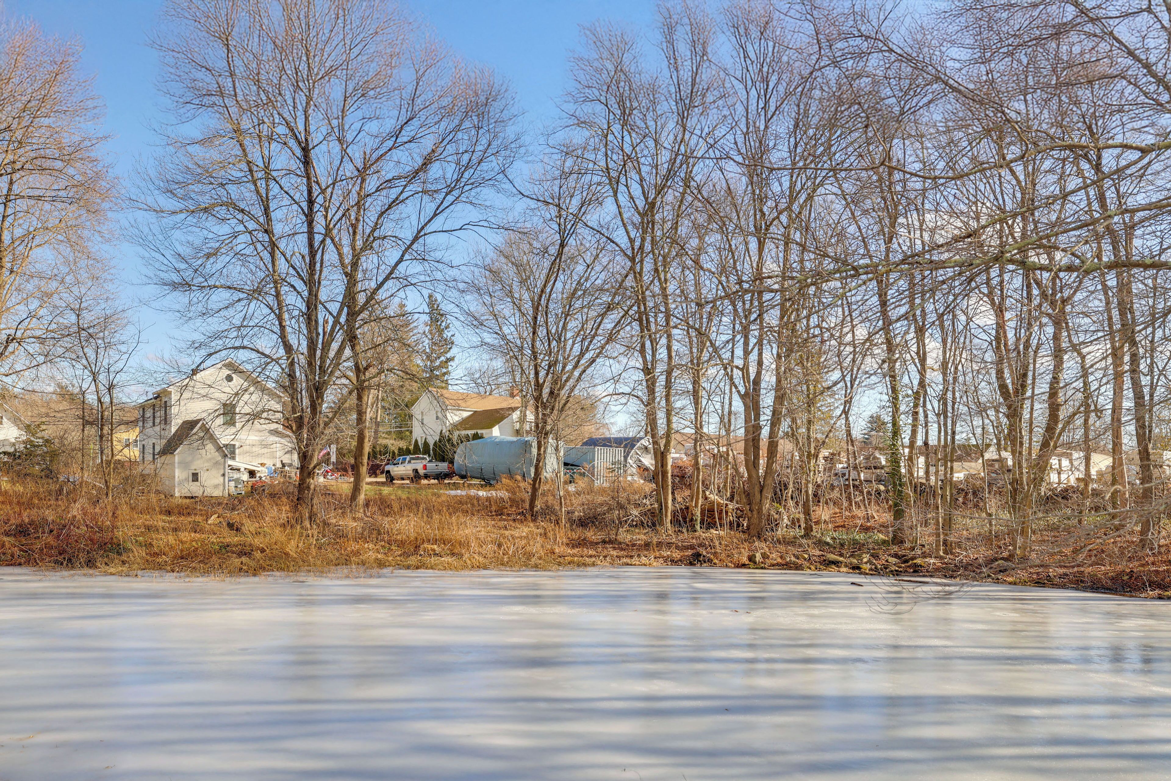 A frozen lake with leafless trees and houses in the background.