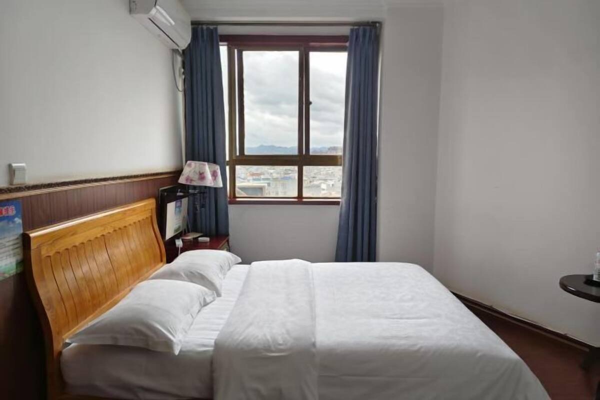 Une chambre d’hôtel avec une tête de lit en bois, un lit avec une literie blanche, une table de chevet avec une lampe, une fenêtre avec des rideaux et une vue sur le paysage urbain.