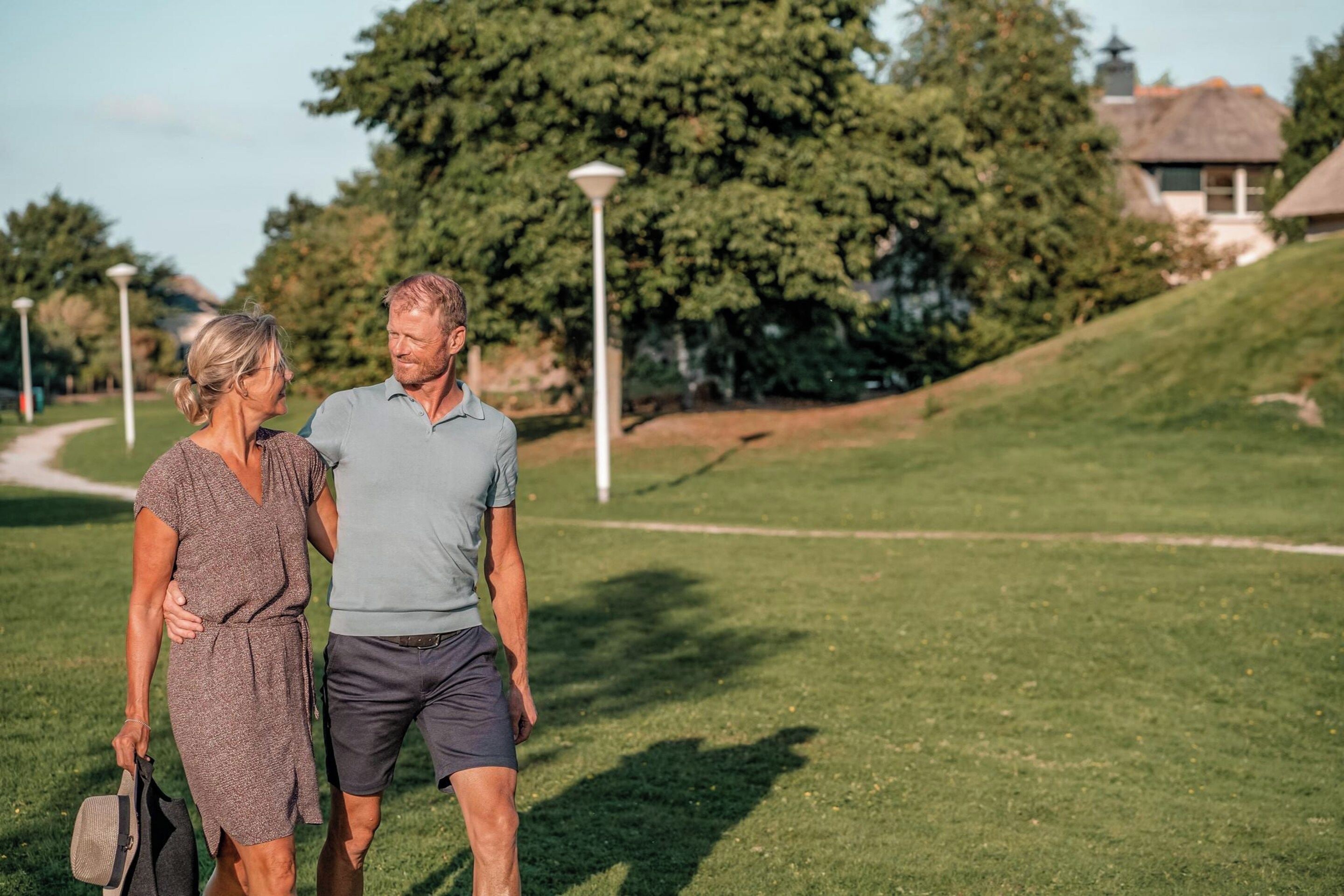 A couple walking on a grassy path with trees and a lamp post in the background.