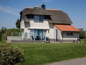 Une maison à deux étages avec un toit de chaume, une véranda à l’avant avec des chaises et un petit jardin.