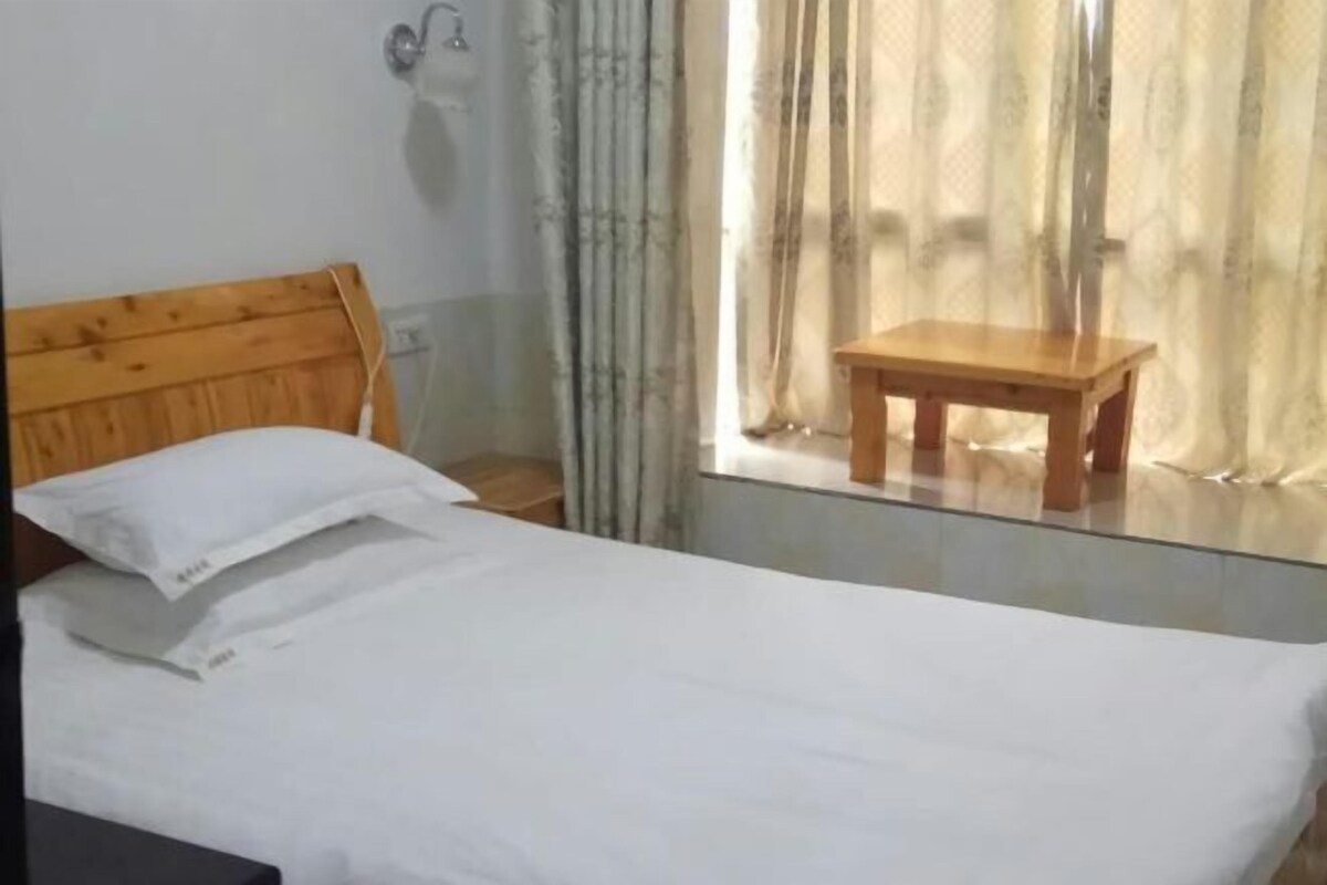 Une pièce avec un lit en bois, du linge de lit blanc, un petit tabouret en bois et une fenêtre avec des rideaux.