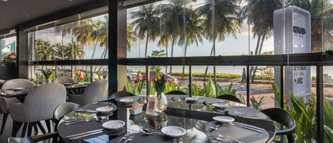 A restaurant with tables set for dining, large windows with a view of palm trees and a body of water.