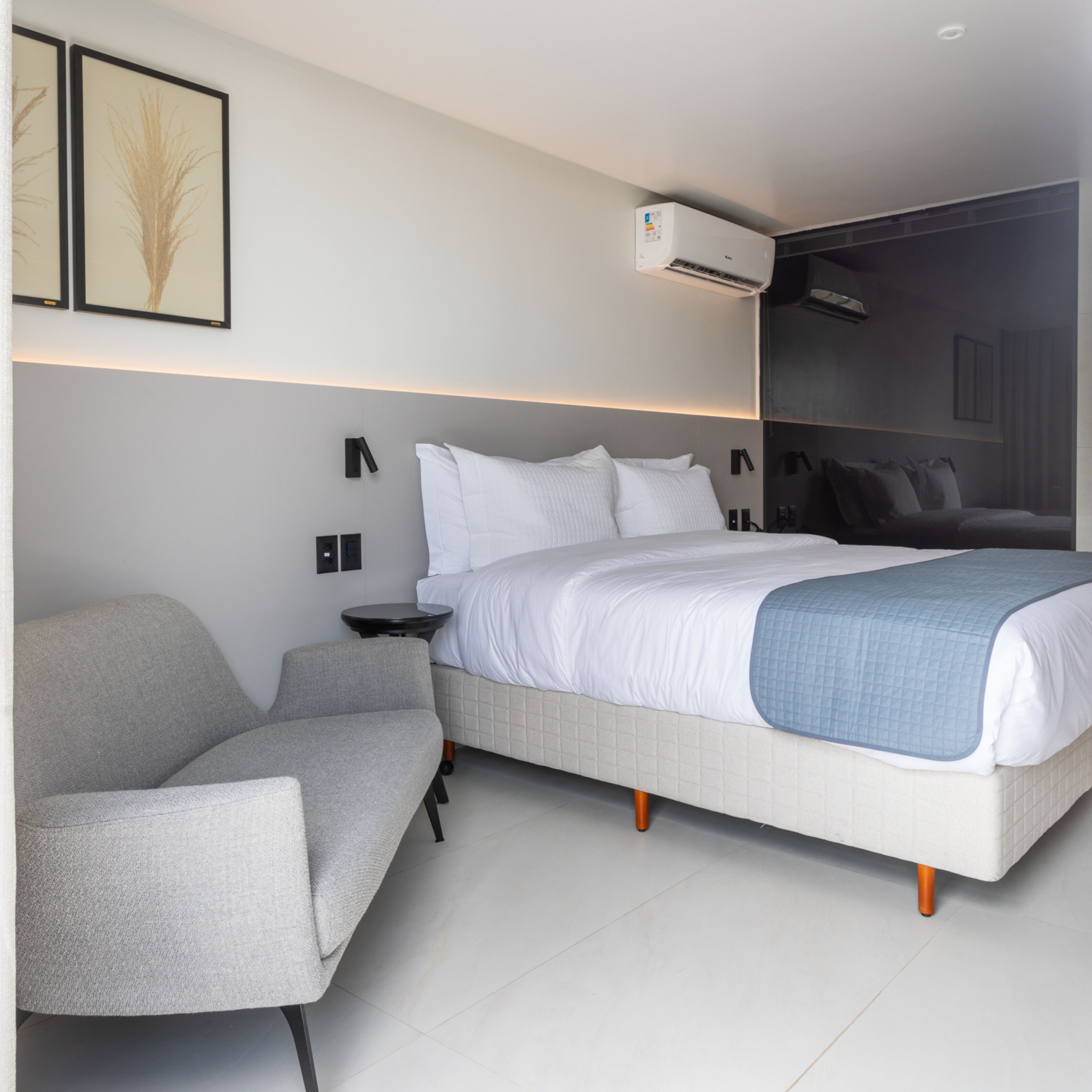 A modern bedroom with a bed, a grey armchair, a bedside table, and an air conditioning unit.