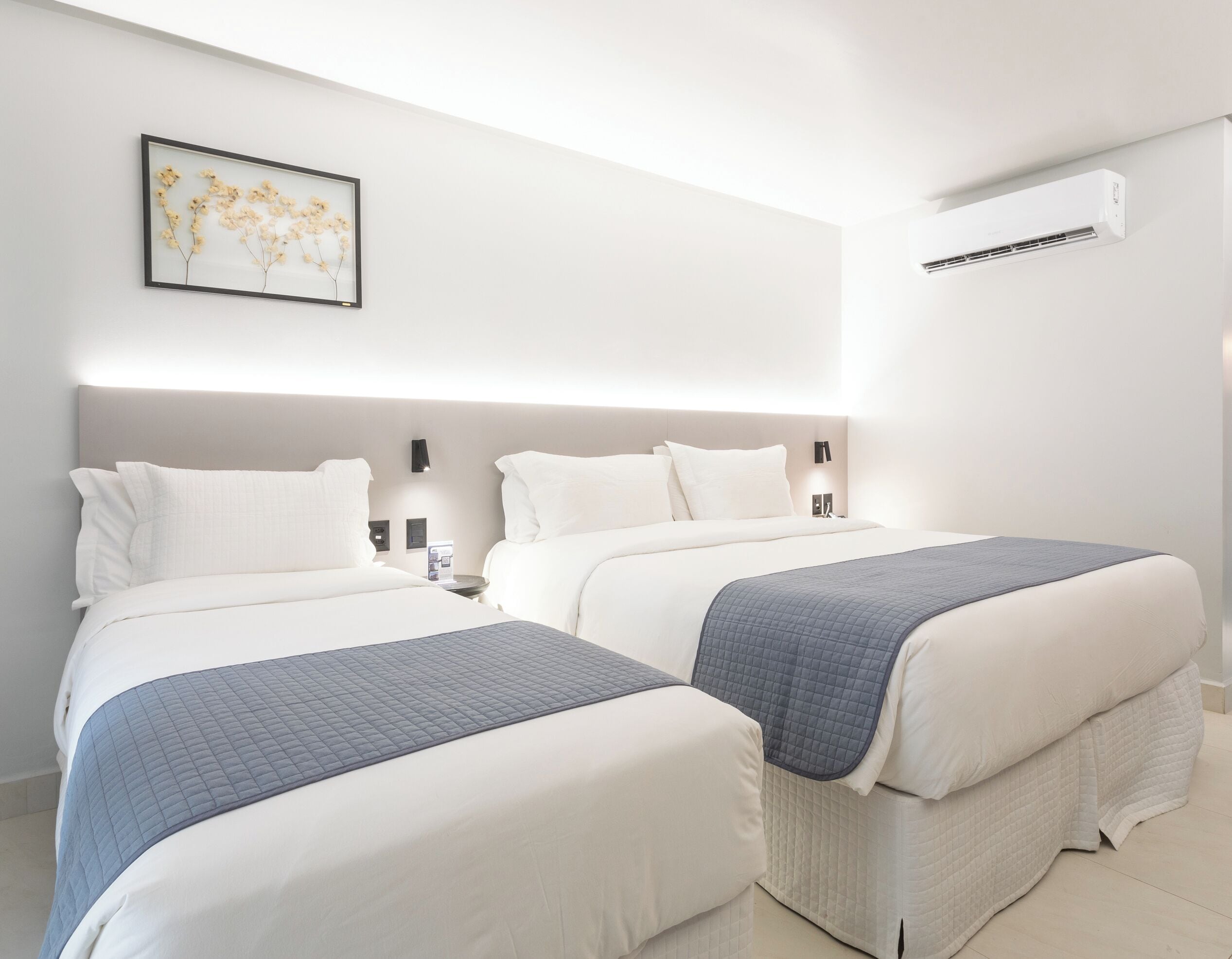 A hotel room with two beds, a framed floral artwork on the wall, and an air conditioning unit.