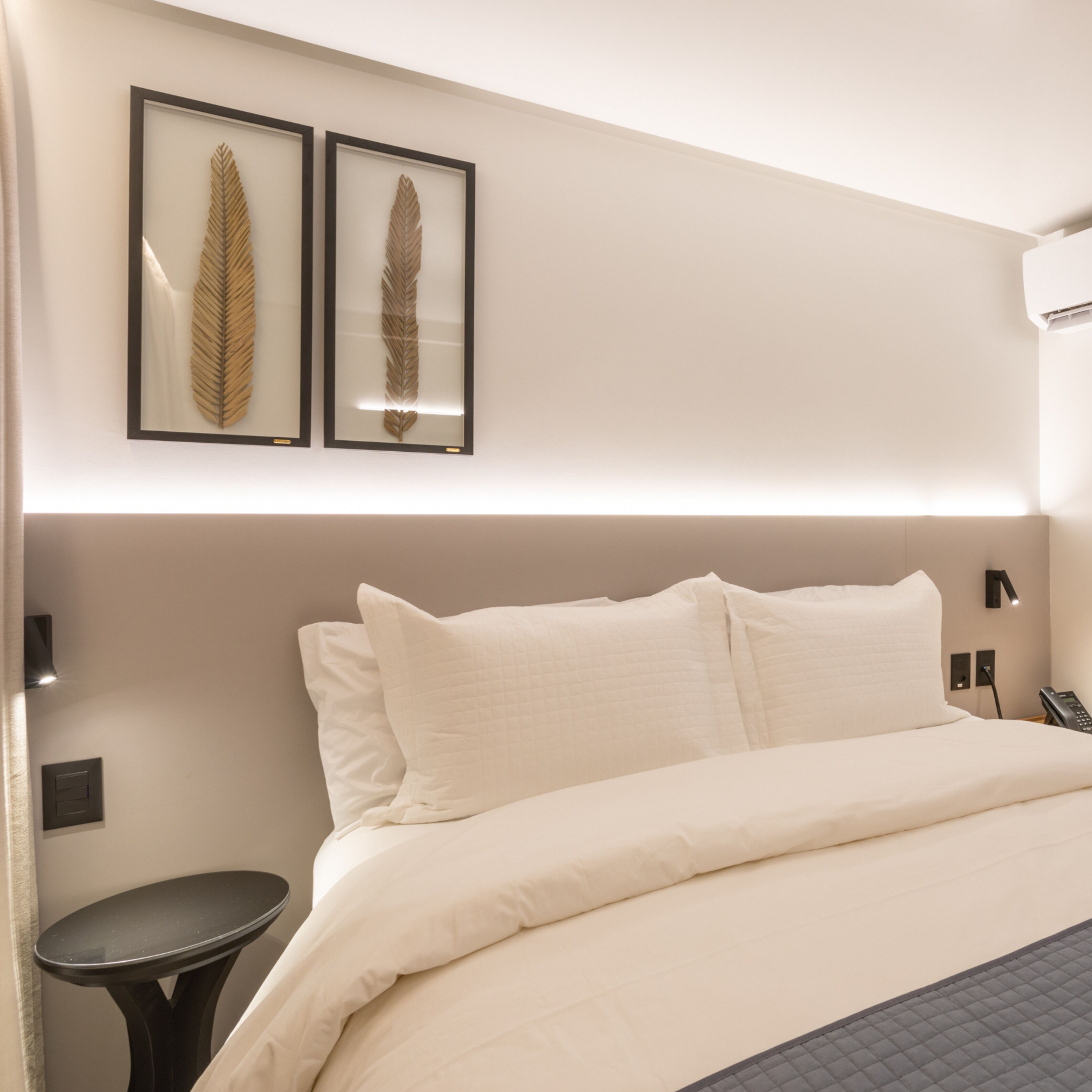 Een hotelkamer met een groot bed, twee lijstjes met bladeren aan de muur, een nachtkastje en een telefoon aan de wand.